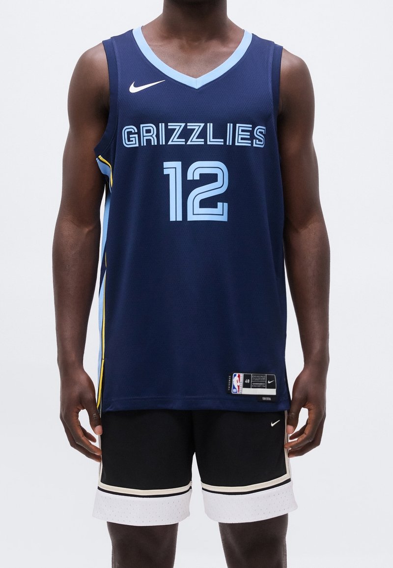 Homme portant un maillot de basketball bleu marine des Memphis Grizzlies avec le numéro 12 et un short noir avec un liseré blanc, debout devant un fond uni.