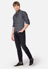 Szary w kratę kamizelka na ciemnej koszuli, w zestawie z przetartymi czarnymi dżinsami i czarnymi sneakersami. Posiada guziki i dopasowany krój.