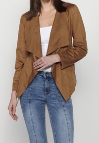 Femme portant une veste marron ouverte sur le devant, un haut blanc et un jean taille haute bleu, une main reposant près du revers de la veste.