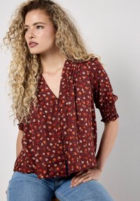 Blusa rossa a fiori con maniche corte e a sbuffo, scollatura a V e chiusura con bottoni. Il tessuto ha una consistenza leggera e morbida con una leggera lucentezza.