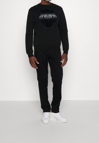 Sudadera negra con un gráfico texturizado de un tigre en la parte frontal, combinada con pantalones negros y zapatillas blancas sobre un fondo neutro.