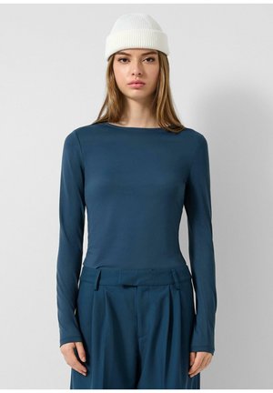 Junge Frau mit langen Haaren, die eine weiße Strickmütze und ein eng anliegendes, langärmeliges Top in Dunkelblau sowie passende, hoch taillierte Hosen trägt.