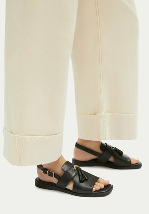 Füße, die schwarze Leder-Sandalen mit offenen Zehen und Quasten tragen, kombiniert mit weiten, cremefarbenen Hosen mit ausgefransten Säumen.