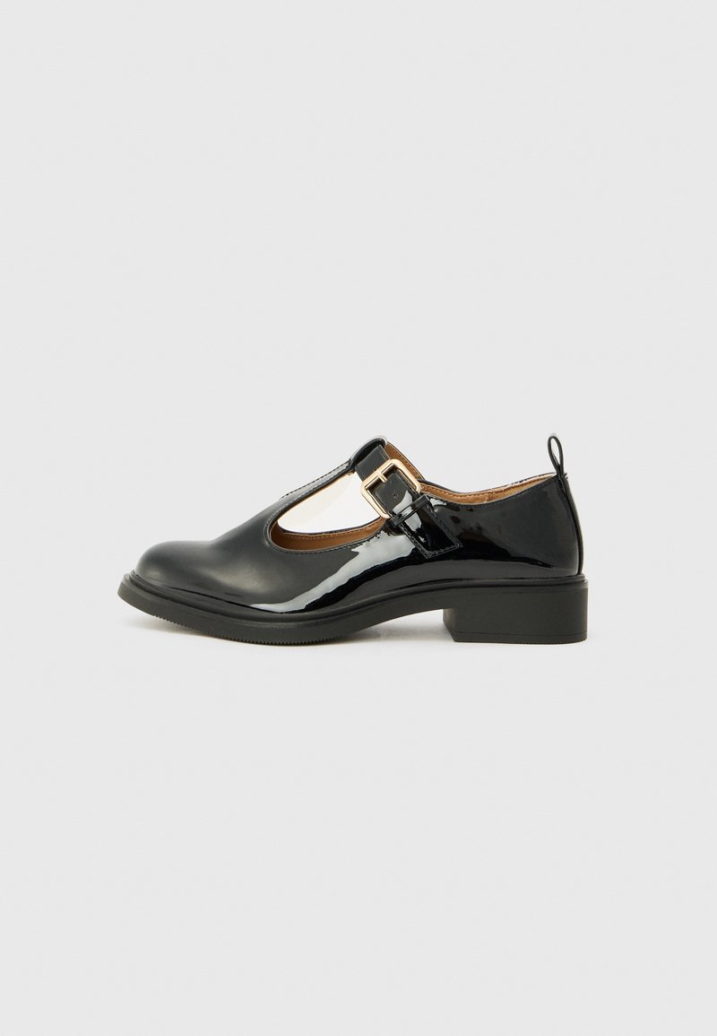 Chaussure Mary Jane en cuir verni noir avec boucle dorée, petit talon et languette à l’arrière sur un fond clair uni.