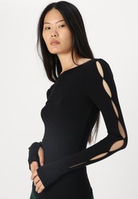 Top negru cu mânecă lungă din material elastic, cu detalii decupate pe brațe și găuri pentru degete. Textură netedă și siluetă ajustată.