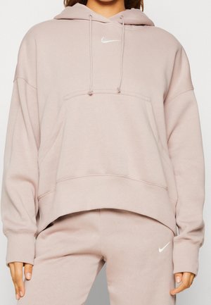 Žena nosí světle růžovou mikinu Nike s kapsou vpředu a odpovídající tepláky s bílými logy Nike swoosh.