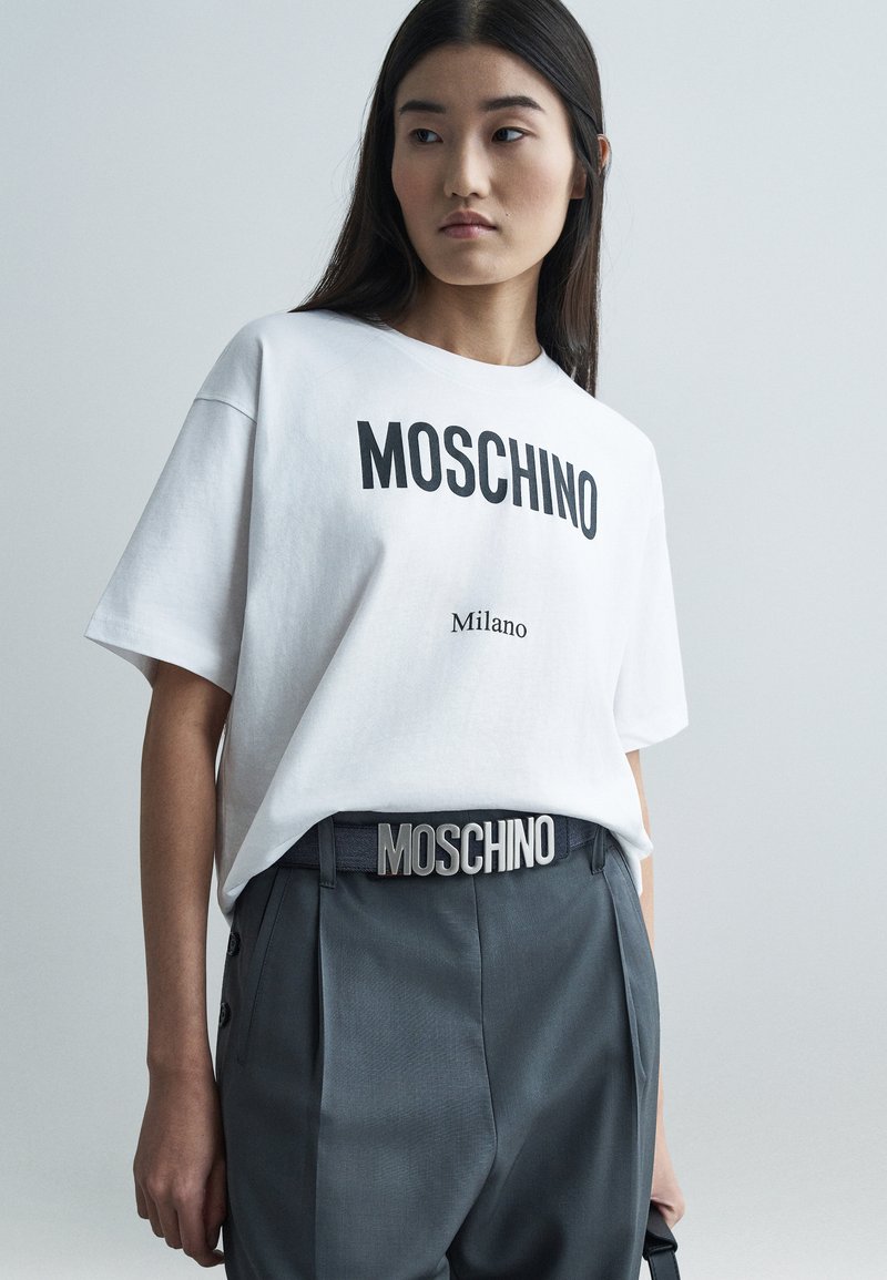 Giovane donna che indossa una maglietta bianca Moschino Milano e pantaloni grigi con una cintura Moschino, in piedi contro uno sfondo chiaro e uniforme.