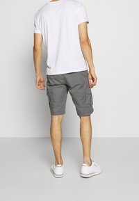 Harmaat cargo-shortsit sivutaskuilla, valmistettu kevyestä kankaasta, yhdistetty valkoiseen T-paitaan ja valkoisiin lenkkareihin, seisomassa yksiväristä taustaa vasten.