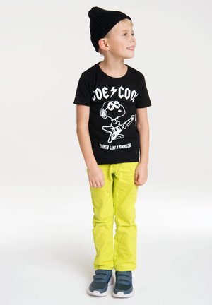 Schwarzes T-Shirt mit weißem Grafik eines Cartoon-Hundes, der Gitarre spielt, kombiniert mit hellgelben Hosen und blauen Sneakers; Junge trägt eine schwarze Mütze.