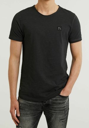 Uomo che indossa una t-shirt nera semplice a maniche corte con un piccolo logo quadrato sul petto sinistro e jeans neri sbiaditi, mano in tasca.