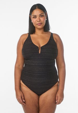 Otse mustade juustega naine kannab musta tekstuuriga üheosalist ujumistrikood sügava V-lõikega, seisab valgel taustal näoga ettepoole.