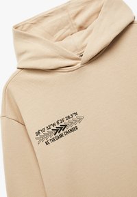 Beige hoodie van gestructureerde stof met zwarte bedrukte tekst en pijl-graphics op de linkerkant van de borst.