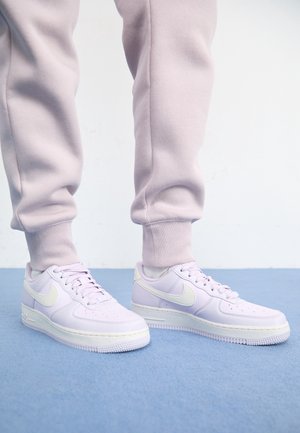 Personne portant un pantalon de survêtement rose clair et des baskets blanches Nike debout sur un sol recouvert de moquette bleue contre un mur blanc.