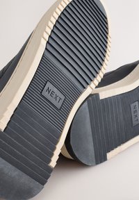 Čierna podrážka z gumy s textúrovaným povrchom, obsahujúca rebrované vzory a béžová medzipodrážka s vyvýrazneným logom "NEXT".