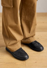 Person wearing loose brown pants and black lace-up shoes standing on wooden floor.