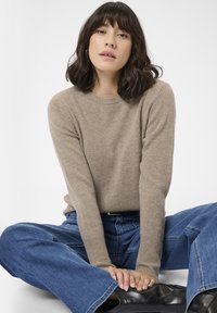 Kvinde med skulderlangt mørkt hår, der sidder med korslagte ben, iført en beige sweater, blå jeans og sorte støvler mod en hvid baggrund.