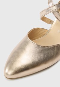 Chaussure dorée métallique avec un bout pointu et un détail de bride croisée. Texture en cuir lisse et doublure intérieure douce.