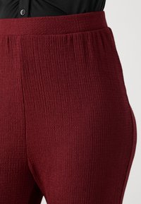 Burgundske rebraste hlače z elastičnim pasom, mehko teksturo in krojem, ki se prilega bokom in stegnum.