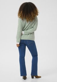 Maglione testurizzato verde chiaro con maniche lunghe, indossato sopra jeans a zampa d'elefante blu. I jeans hanno tasche posteriori con cuciture a contrasto. Stivaletti con tacco marroni.