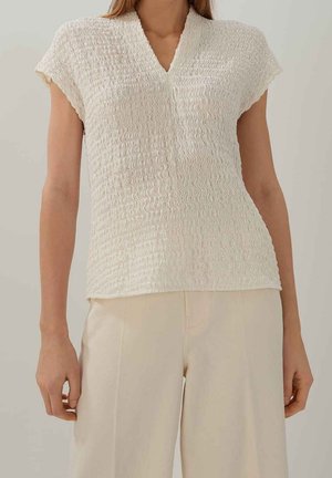 Krémově zbarvený top s texturovanou, vlnitou látkou a výstřihem do V. Krátké rukávy a přiléhavý střih, kombinovaný se světle zbarvenými kalhotami.