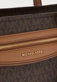 Κοντινή λήψη τμήματος καφέ τσάντας Michael Kors με χρυστό φερμουάρ και λογότυπο σε υφασμένο ύφασμα με επαναλαμβανόμενο μοτίβο MK.