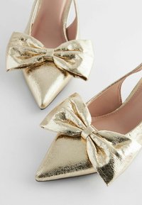 Kultaiset metallinhohtoiset slingback-korkokenkiä, joissa on terävät varpaat ja suuret, teksturoidut soljet edessä. Sujuva sisus ja kapeat hihnat.