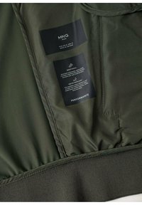 Wnętrze oliwkowej kurtki z naszywkami z tkaniny informującymi o "oddychalności" i "wodoodporności". Posiada elastyczny ściągacz i gładką podszewkę.