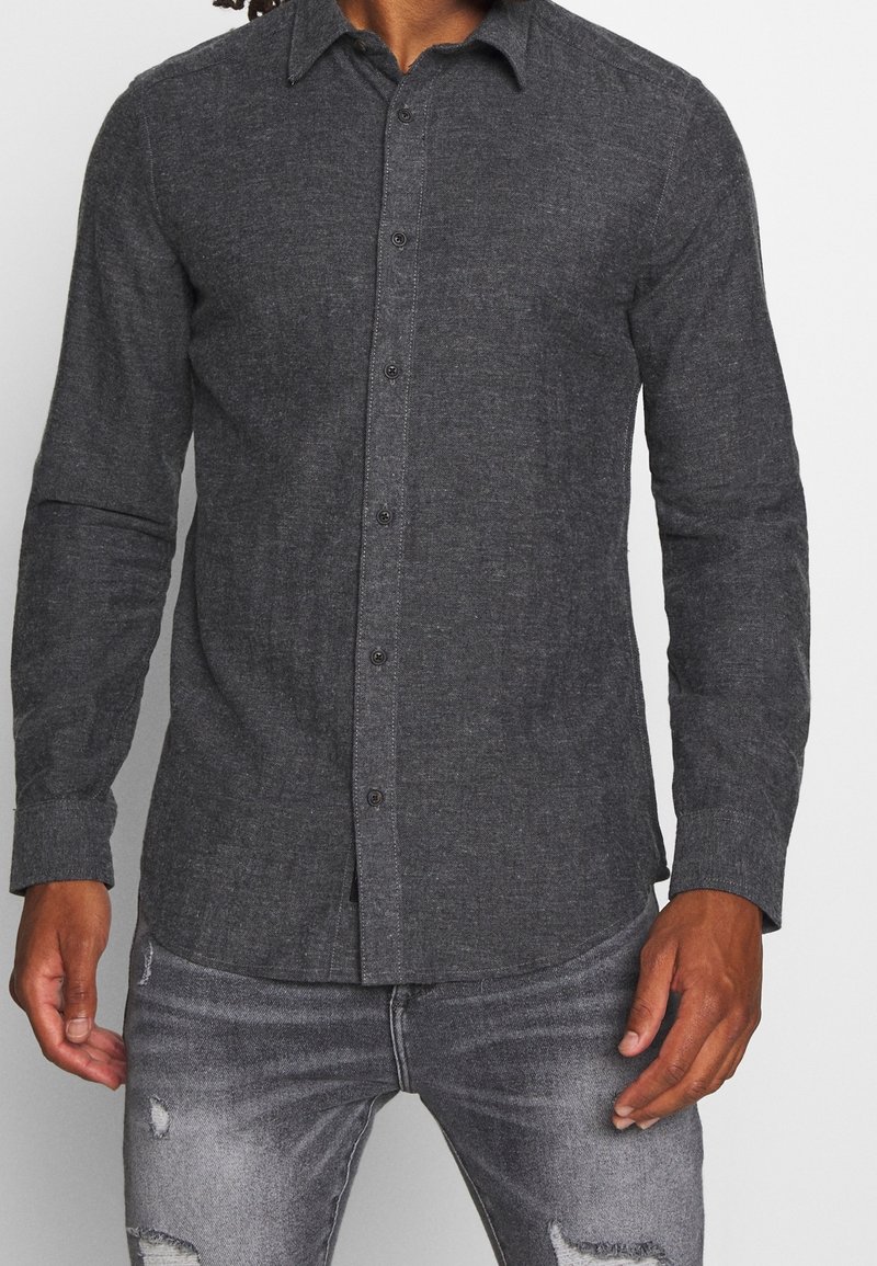 Uomo che indossa una camicia grigia scura a maniche lunghe con bottoni abbinata a jeans neri strappati e scoloriti, in piedi contro uno sfondo semplice.