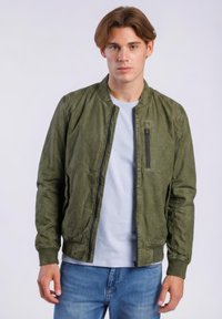 Nuori mies vaaleanruskein hiuksin, pukeutuneena vihreään vetoketjulliseen takkiin valkoisen t-paidan ja sinisten farkkujen päällä, seisomassa neutraalia taustaa vasten.