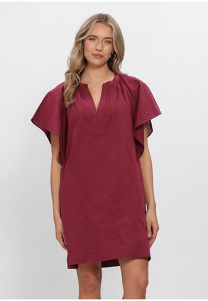 Vrouw met lang blond haar die een losvallende, korte mouwen, bordeauxrode jurk met een gespleten hals draagt, staand tegen een effen witte achtergrond.