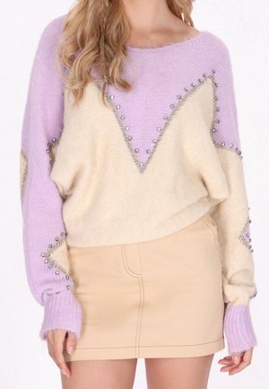 Femme portant un pull duveteux lilas et crème avec des détails en perles, assorti à une mini-jupe beige sur un fond uni.