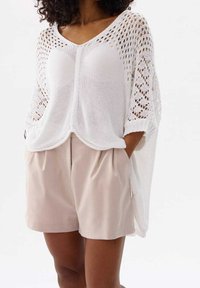 Weißer gestrickter Poncho mit V-Ausschnitt, Lochmuster und kurzen Ärmeln, kombiniert mit beigen Shorts mit Falten und Taschen.