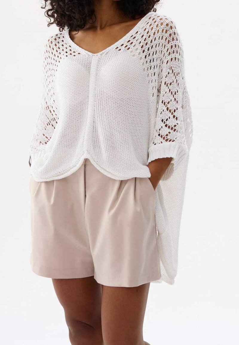Weißer gestrickter Poncho mit V-Ausschnitt, Lochmuster und kurzen Ärmeln, kombiniert mit beigen Shorts mit Falten und Taschen.
