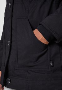 Svart ytterjacka med en synlig bröstficka, texturerad tyg och ribbad manschettdetalj. Tryckknappsstängning vid fållen.