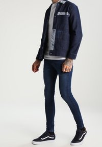 Marinblå denimjacka med randiga detaljer, knäppning med knappar och två framfickor, i kombination med mörkblå slim-fit jeans och svarta sneakers.
