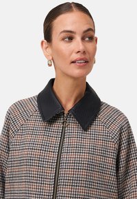 Veste à motif pied-de-poule en gris et marron avec un col en cuir noir et une fermeture éclair à l'avant. Elle présente un tissu texturé et audacieux ainsi que des manches raglan.