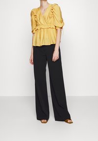 Blusa gialla arricciata con scollatura a V e vita cinchata, abbinata a pantaloni neri a gamba larga e sandali con cinturino marroni.