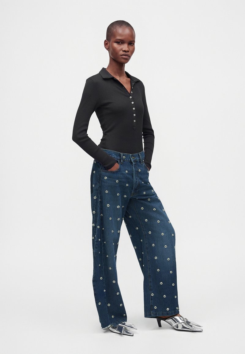 Femme portant une chemise noire à manches longues boutonnée, un jean bleu large avec des détails œillets, et des talons métalliques pointus, debout les mains dans les poches.