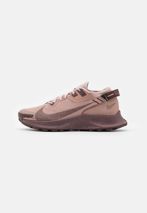 Jasnoróżowy i brązowy but do biegania Nike z teksturowaną podeszwą, siateczkową cholewką i oznakowaną pętelką na białym tle.