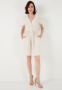 Short-sleeved, cream dress with a v-neckline, button front, gathered waist, and pockets, made from textured fabric.