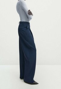 Pantalon large bleu marine en tissu lisse, doté d'une taille haute et de plis nets, associé à des chaussures noires élégantes à bouts pointus.