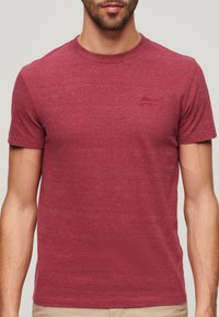 Camiseta de manga corta de color rojo oscuro hecha de tejido texturizado. Presenta un pequeño logo bordado en el lado izquierdo del pecho. Cuello clásico de estilo crew.