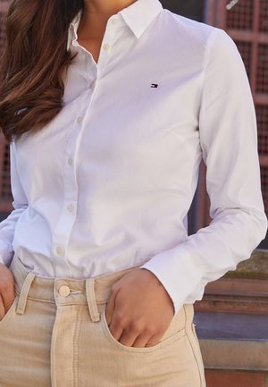 Mujer que lleva una camisa blanca ajustada abotonada con un pequeño logo en el pecho y pantalones de tiro alto beige claro, manos en los bolsillos.