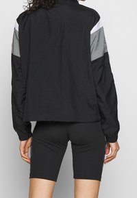 Svart vindjacka med grå och vita detaljer, långa ärmar, avslappnad passform och textilmaterial. Svarta högmidjade cykelshorts.