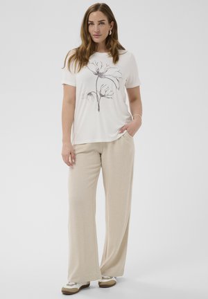 Mujer con cabello castaño que lleva una camiseta blanca con estampado floral negro, pantalones beige de pierna ancha y zapatillas blancas, de pie con una mano en el bolsillo.