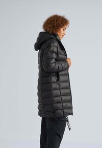 Parka nera con cappuccio, caratterizzata da un design trapuntato e tasca laterale con zip. Include un'etichetta con il nome del marchio "JACKIT" sul lato.