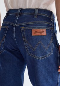 Person iført mørkeblå Wrangler-jeans med synlig syning på baglommen og lædermærke med brandlogo på højre lomme.