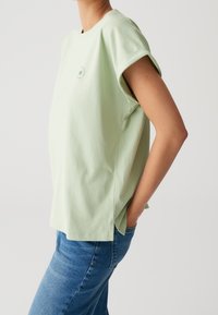 T-shirt di cotone verde chiaro con maniche corte arrotolate, scollo rotondo, fessure laterali e una piccola stampa grafica sul davanti. Indossato con jeans blu.