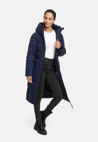 Lang marineblauwe pufferjas met een capuchon, voorzien van doorgestikte naden en zijzakken, gecombineerd met zwarte jeans en enkellaarsjes.