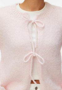 Gros plan sur une personne portant un cardigan tricoté rose doux noué avec deux nœuds sur un haut blanc côtelé et un pantalon blanc.